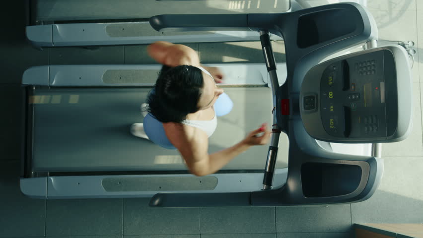 Well-built young woman in white top and sneakers, blue leggings, with long brown hair is running on treadmill. Top view. The monitor shows results. High quality 4k footage Royalty-Free Stock Footage #1103075621
