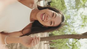 Smiling brunette girl dressed in white top and jeans making video call from mobile phone while standing in city park, Young woman waving hand