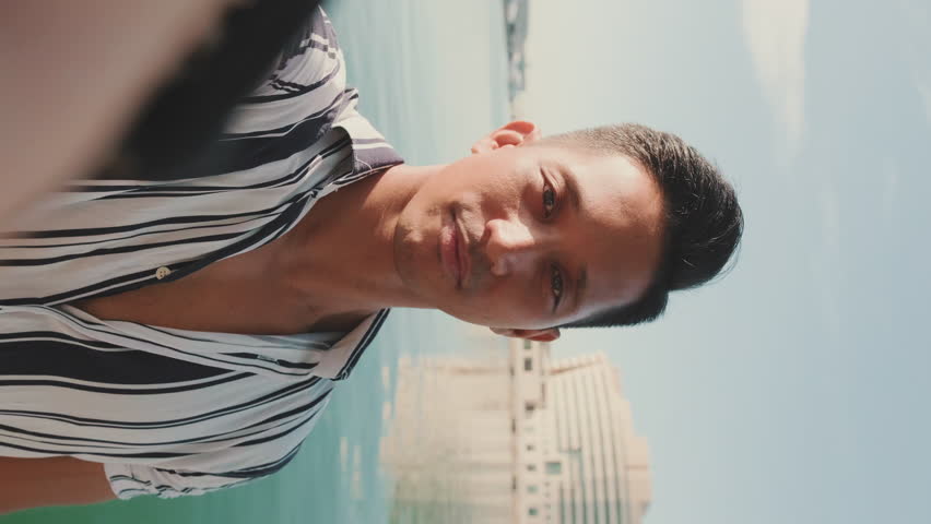 VERTICAL VIDEO: Close-up of young man standing on dock taking selfie with cell phone