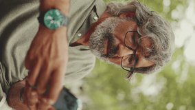 VERTICAL VIDEO: Positive middle-aged man with gray hair and beard wearing casual clothes sits on bench and uses mobile phone. Mature gentleman in eyeglasses writes message on smartphone