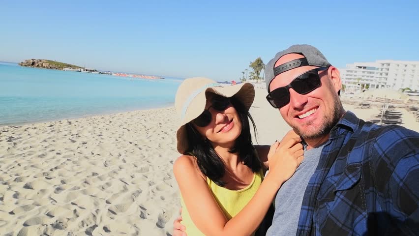 Happy multiracial couple taking selfie on summer vacation - Trendy man and woman having fun on Cyprus Nissi beach. Friendship and holidays concept with guy and girl enjoying summer