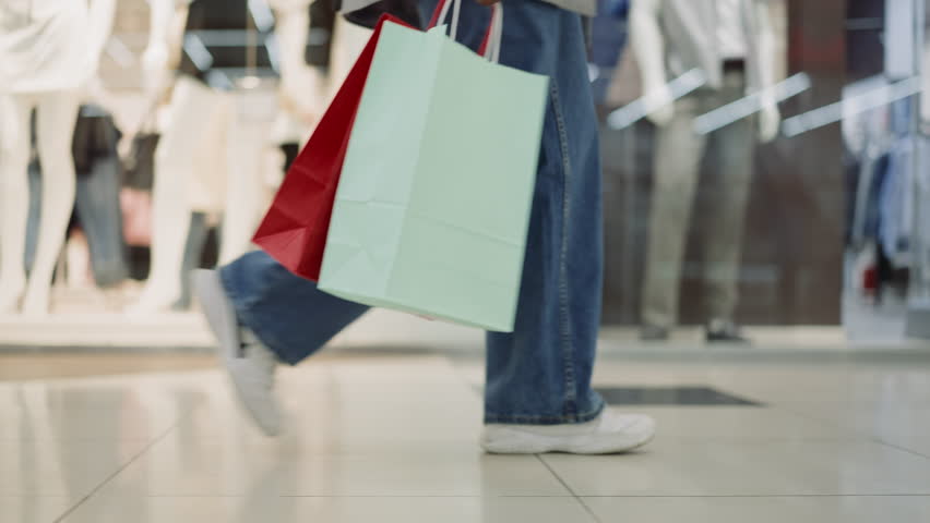 Woman legs in jeans and sneakers walking across mall closeup slow motion. Lady customer holds shopping bags after visiting fashion stores. Hobby and leisure