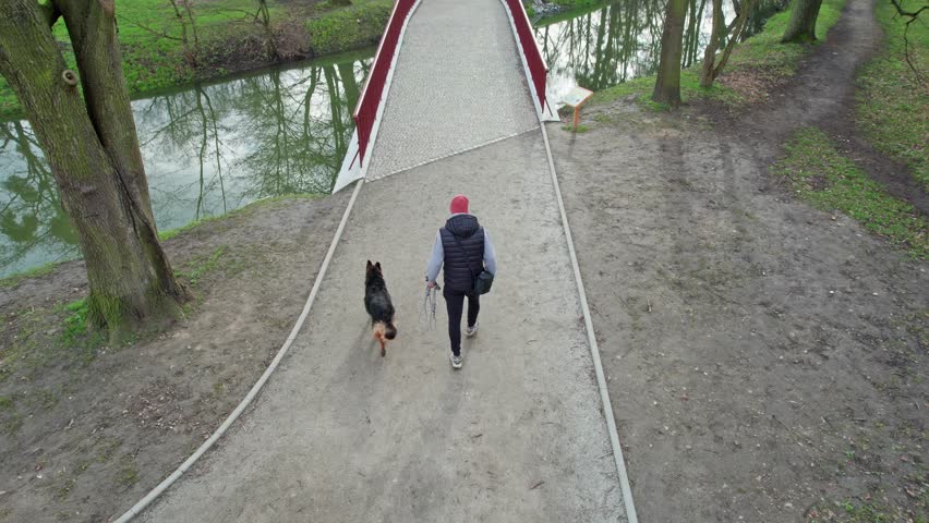 A man with his dog takes a walk in the park on a spring day, wearing a cap and a vest, the sky is cloudy.