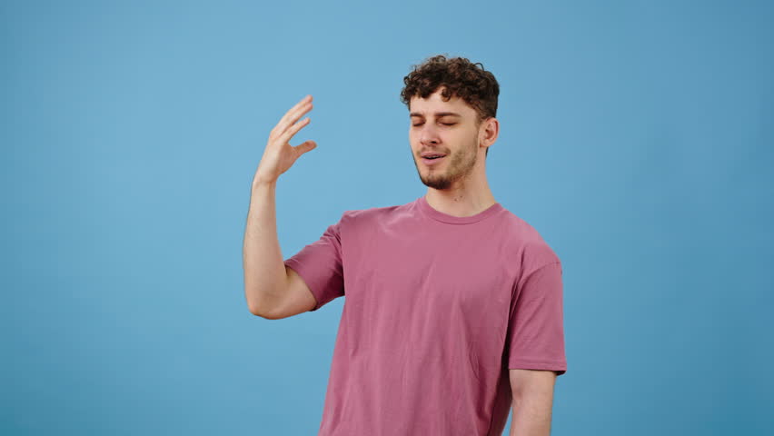 Caucasian young curly haired man in pink t-shirt, showing tasty gesture, kisses fingers and says bellissimo, isolated on blue background. Gourmet culinary concept