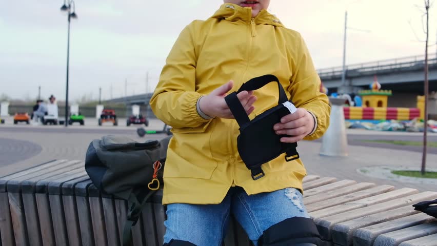A boy in a yellow jacket puts on elbow pads on his hands on a bench outside before riding a scooter.