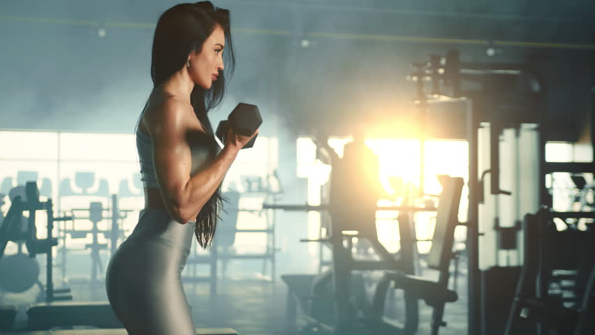 Medium long shot of athletic woman profile doing dumbbell hammer curl in gym. Slow motion. Misty fully equipped gym with sun rays in the background. High quality 4k footage Royalty-Free Stock Footage #1103088151