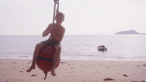 A video showing a boy looking far away while swinging on a buoy boll on rope on the beach during daytime in Thailand