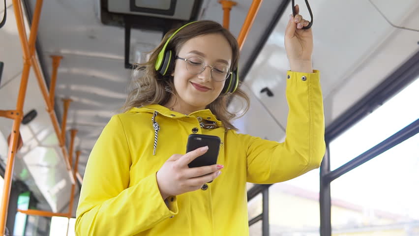 Smiling young woman wearing headphones standing on a bus listening to music and using smartphone.. Royalty-Free Stock Footage #1103094761