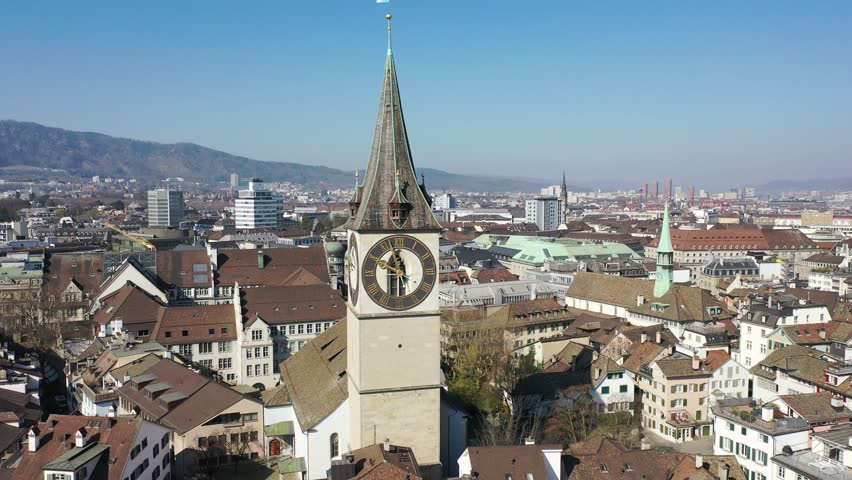 Aerial view of Zurich, Switzerland, showcasing the iconic St. Peter’s Church clock tower and historic architecture under a clear blue sky. Peaceful cityscape with lake and mountains in the background.