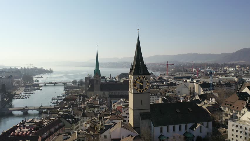 Aerial view of Zurich, Switzerland, showcasing the iconic St. Peter’s Church clock tower and historic architecture under a clear blue sky. Peaceful cityscape with lake and mountains in the background.