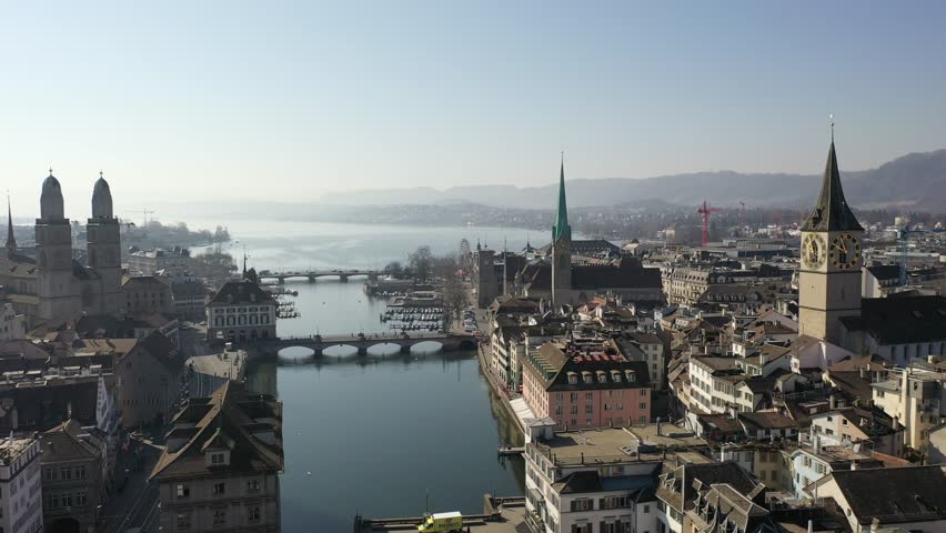 Drone view of Zurich’s old town with Limmat River, traditional houses and church spires under clear sky. Beautiful Swiss cityscape blending history, nature, and urban life.