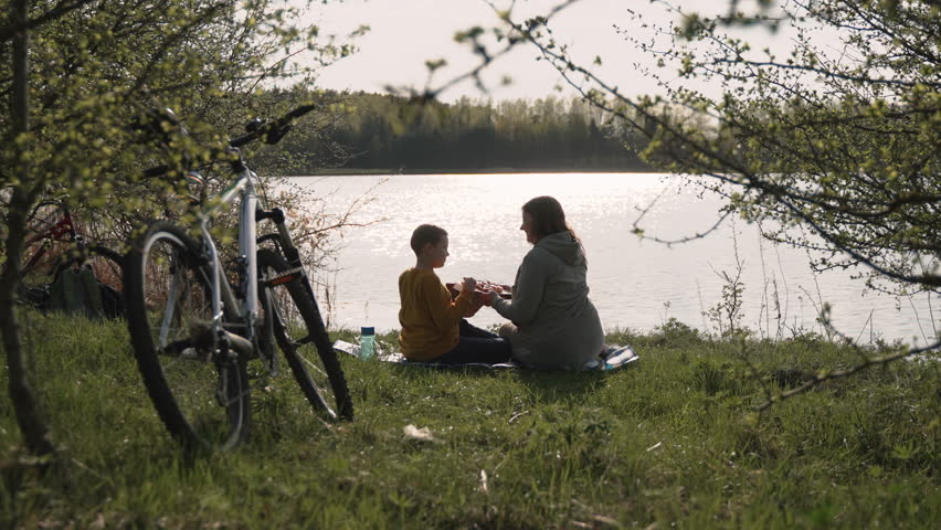Son shows mother appetizing slice of pizza at picnic in park, back view. Happy caucasian family is resting on shore of lake after bike ride. Sunlight reflects off the surface of pond.