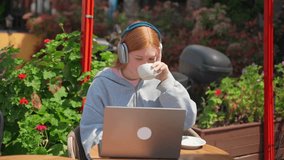 Teenage girl headphones using laptop in cafe outdoor drink coffee. Cheerful young woman sitting watching video listening to music phone student working essay, exam study in flower background