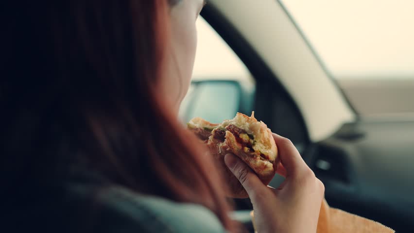 Young hungry woman eats roll with meatball while sitting in car. Teenager travels, having snack on road without leaving car. Girl passenger eats burger in car. Delicious food bun with cutlet. Tourist Royalty-Free Stock Footage #1103113259