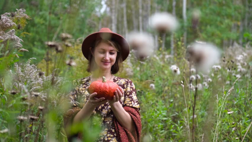 Smiling woman holding an orange pumpkin in her hands against the backdrop of an autumn forest. The concept of autumn comfort, fading nature, Thanksgiving