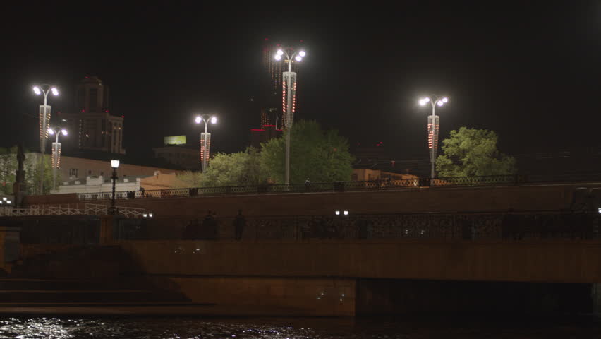 Evening city. Stock footage. A dark view of the city with a river over which there is a bridge with a road for tourists and new buildings surrounded by lanterns.