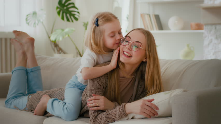 Happy motherhood family bonding affectionate love Caucasian mother lying on couch sofa daughter little girl child piggyback kiss mom kissing lovely parent warm communication talking sweet tenderness Royalty-Free Stock Footage #1103117347