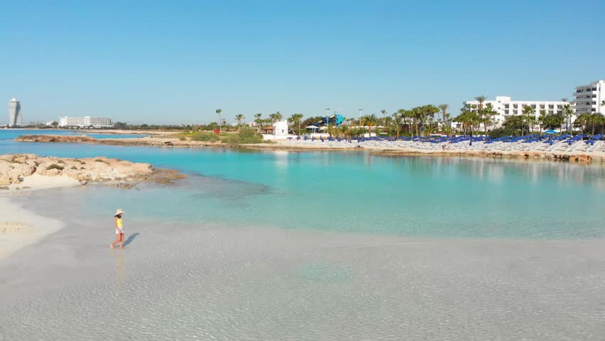 Aerial view woman tourist walk on beautiful Nissi beach in Cyprus. Famous travel visit destination. Popular famous white sand beach. Solo traveler visit Nissi island beach