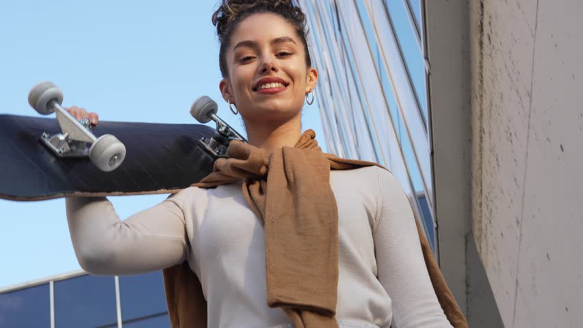 One woman young adult caucasian female stand outdoor posing portrait happy confident in front of white wall modern building smile slow motion hold skateboard