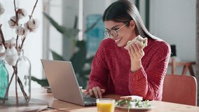 Video of beautiful young woman working with laptop while having healthy sandwich for breakfast at home