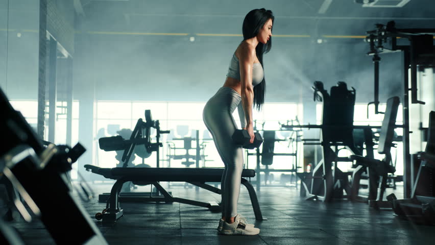 Full shot of athletic young cute woman performing dumbbell Romanian deadlift. Profile. Slow motion. Fully equipped misty gym, sun rays in the background. High quality 4k footage Royalty-Free Stock Footage #1103141559