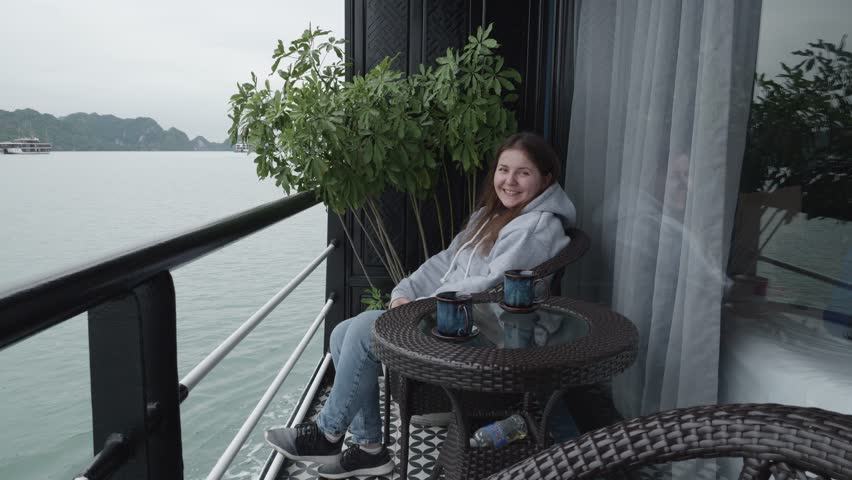 Woman in her 30s Sitting On The Balcony Of A Cruise Ship. Smiling At Camera While Navigating On The Waters Of Halong Bay In Vietnam. wide