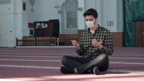 Young man with mask on his face due to pandemic sitting in mosque praying with open hands, muslim youth praying in masjid