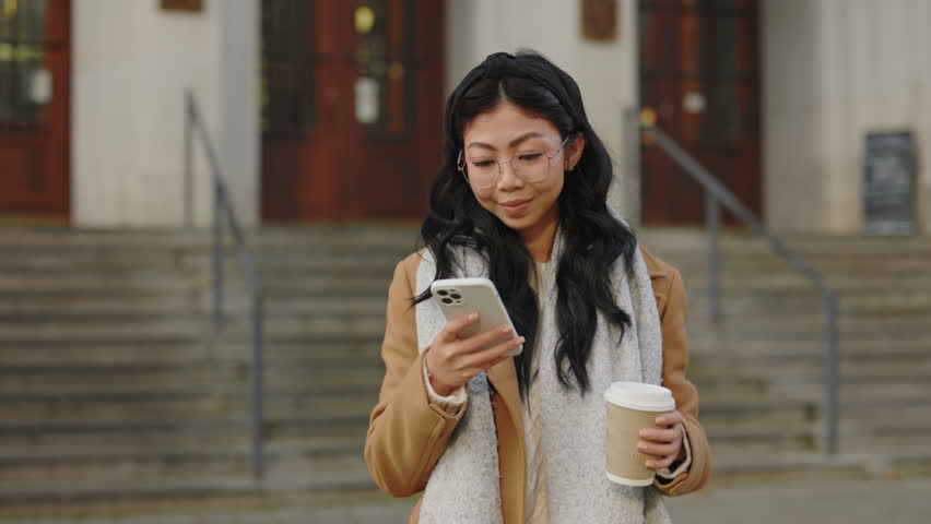 Smiling Asian Woman Using Smartphone and Surfing the Internet while Holding a Takeaway Coffee Cup on the Street. Communication and Technology Concept Royalty-Free Stock Footage #1103157585