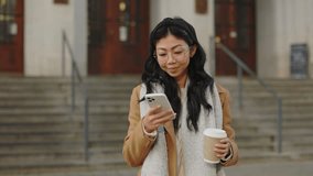 Smiling Asian Woman Using Smartphone and Surfing the Internet while Holding a Takeaway Coffee Cup on the Street. Communication and Technology Concept - Powered by Shutterstock - Get 15% off with code: PIKWIZARD15