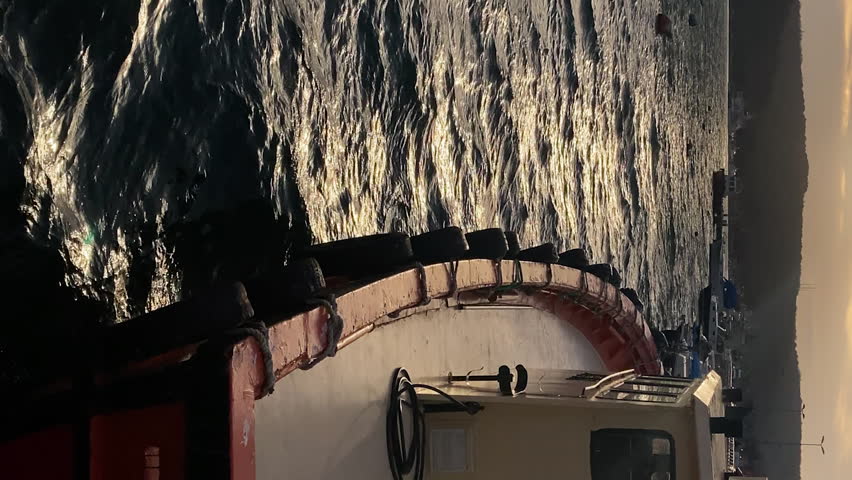 VERTICAL: View of the waterfront at sunset with red trawler tied to the shore and swaying on the waves. Beautiful highlights on the water. FullHD footage