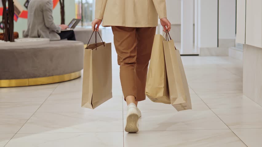 Back view of woman shopaholic is holding shopping bags and strolling around the shopping mall. Satisfied customer walks through the hall of beautiful shopping center, passing man who using laptop. Royalty-Free Stock Footage #1103160473