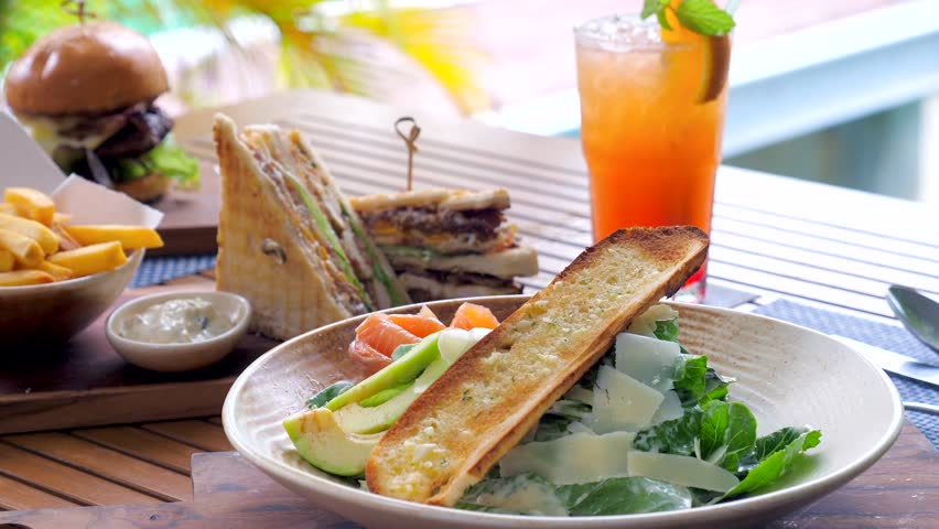 Delicious lunch served on a wooden table in an outdoor cafe with a variety of food including salad, sandwiches, burger, and french fries with a tropical drink. Tasty food on holiday or vacation.