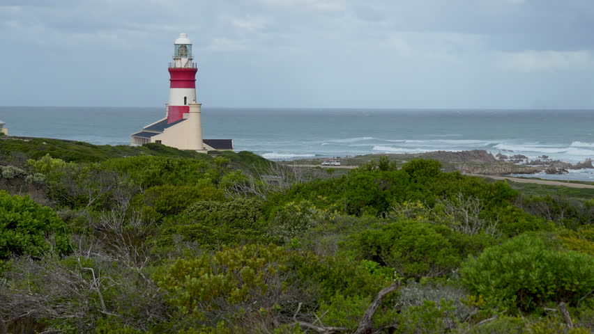 Rich green fynbos covered dunes, an historic red and white striped lighthouse on a grey cloudy day at Cape Agulhas in South Africa.