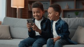 Children gadget addiction two happy laughing African American little boys brothers browsing mobile phone watching cartoons online without parental control at home couch kids siblings using smartphone