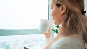 happy blonde woman holding cup of hot drink, drinks coffee enjoy aroma looking at window, top view city winter sky snow falls. Cozy white room. Blonde hair back. Girl fashion model smiling face. 4k - Powered by Shutterstock - Get 15% off with code: PIKWIZARD15