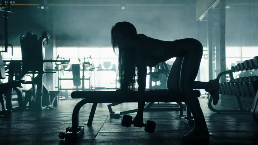 Black female silhouette with fit body, long hair performing one arm dumbbell row in gym. Cardio area, large window, big mirror in dark misty background. High quality 4k footage