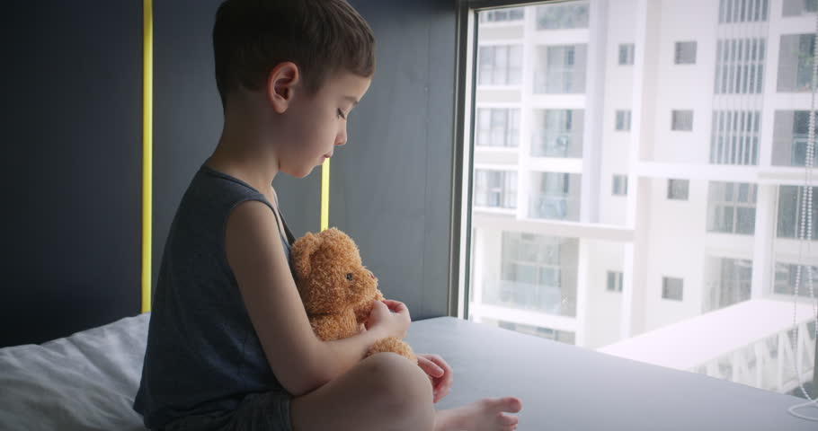 Little boy upset child of preschool age sits near window on his bed, holds his plush bear in his hands,looks out window into street.Violence against children in family.Sad kid.Child's fear loneliness. Royalty-Free Stock Footage #1103179651