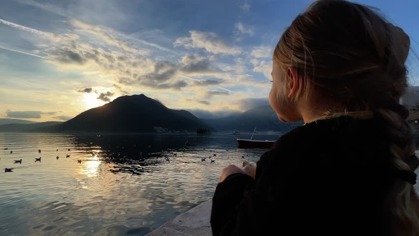 Little girl eats bread on the pier and looks at the seagulls floating on the sea at sunset