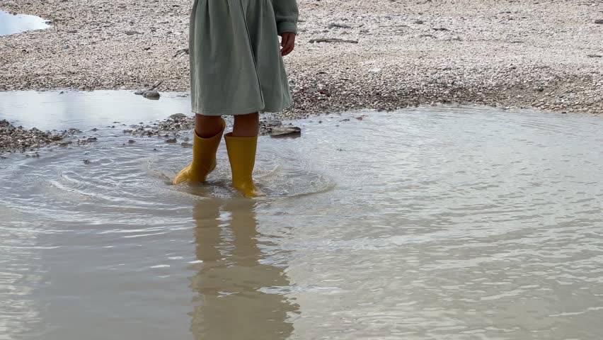 Little girl in rubber boots walks through a puddle on the seashore