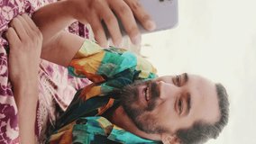 VERTICAL VIDEO: Young happy man talking by video link on mobile phone on the beach