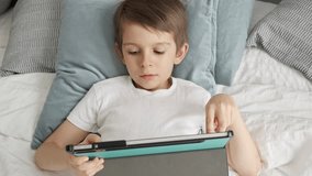 A little boy is lying on his bed and playing with a tablet computer during the day. Child using gadget, education and development