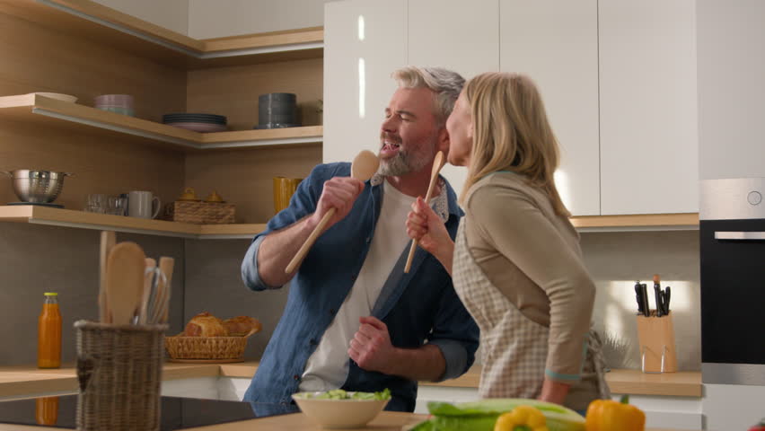 Middle-aged couple happy family cheerful wife and husband singing song having fun fooling around at kitchen together cook fresh vegetables salad adult woman and man sing in kitchenware utensil spoons Royalty-Free Stock Footage #1103200221
