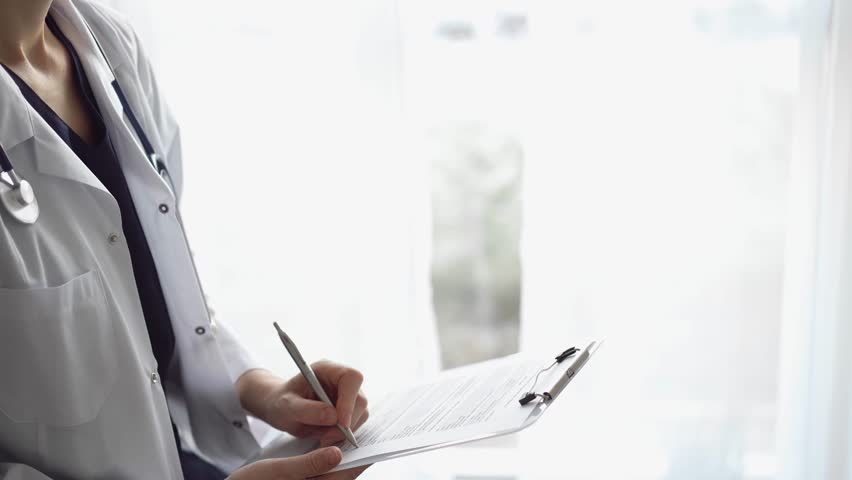 Doctor woman at work using a white clipboard while writing medical notes. An unknown physician standing near the panorama window in hospital. Medicine Royalty-Free Stock Footage #1103205589
