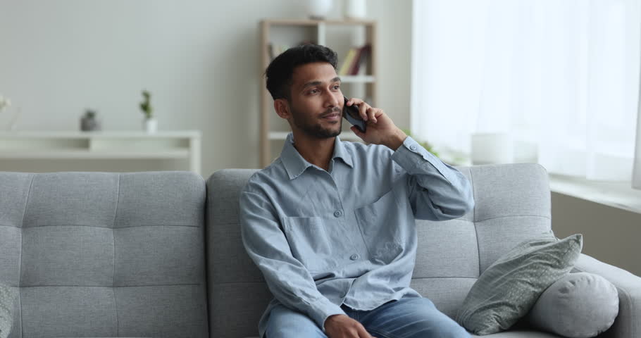 Indian man blab on cell phone, sit on couch holds modern gadget, having informal conversation, lead pleasant talk to friend, arrange meeting, share news spend time at home enjoy carefree communication Royalty-Free Stock Footage #1103218533