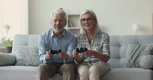 Happy elderly spouses spending family weekend at home sitting on couch in living room, use modern technology, have fun together playing computer games with joypad, carefree couple enjoy competitions