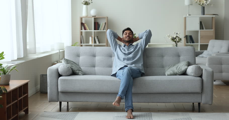 Carefree Indian guy puts hands behind head relaxing seated on soft, comfortable couch in fashionable, light living room, smile enjoy fresh air indoors. Stress-free leisure, daytime rest at smart home Royalty-Free Stock Footage #1103218655