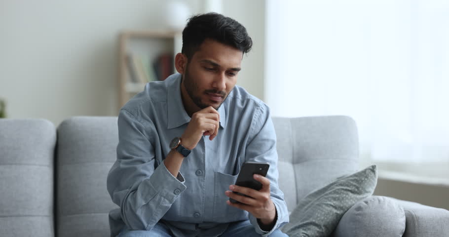 Focused young Indian guy spend free time at home sit on sofa in living room, use mobile application on smartphone, browse web, search useful information, read or write e-mail, share news, chat on-line Royalty-Free Stock Footage #1103218711