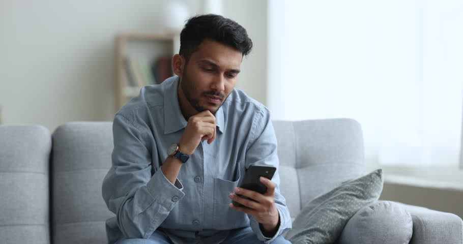 Focused young Indian guy spend free time at home sit on sofa in living room, use mobile application on smartphone, browse web, search useful information, read or write e-mail, share news, chat on-line Royalty-Free Stock Footage #1103218711
