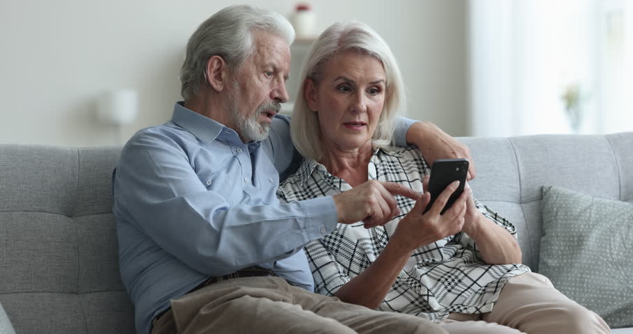 Overjoyed aged wife and husband sit on sofa in living room, use online application on mobile phone, receive good news, amazed by win in interactive on-line game, get prize in web lottery, feel happy Royalty-Free Stock Footage #1103218773