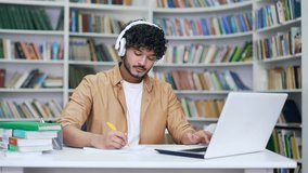 Young college student wearing headphones studying while watching online webinar, class webcast, looking at laptop, e-learning, taking notes or remote teacher video call, prepares for university exams